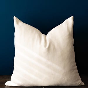 May include: A large, off-white square pillow with a textured surface. The pillow is centered against a dark blue background and rests on a wooden surface. The pillow's shape is defined by its pointed corners.