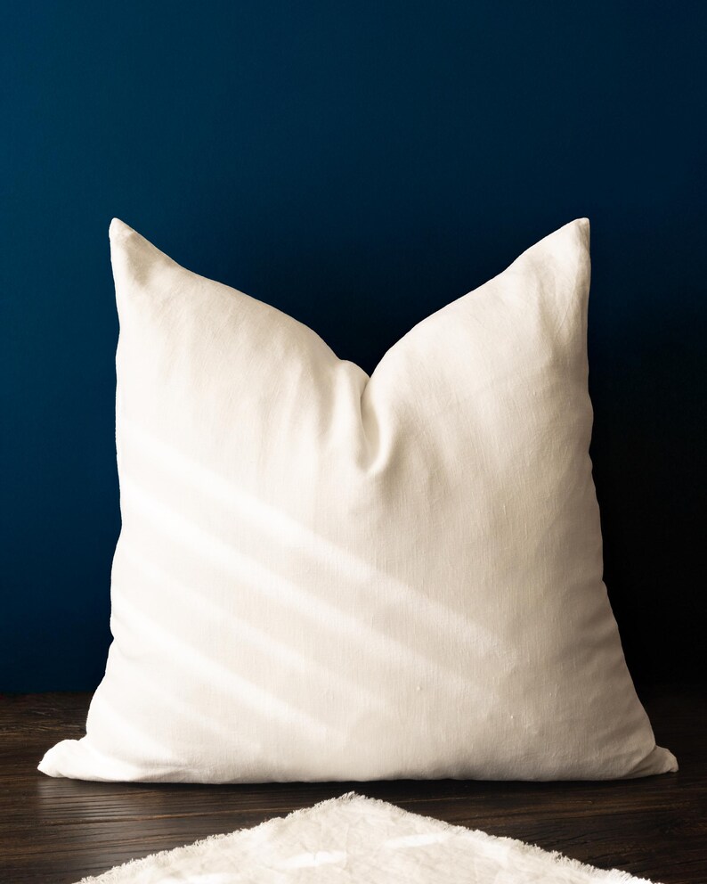 May include: A large, off-white square pillow with a textured surface. The pillow is centered against a dark blue background and rests on a wooden surface. The pillow's shape is defined by its pointed corners.