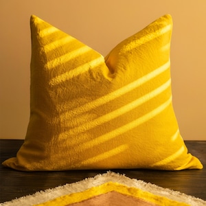 May include: A bright yellow decorative pillow with a textured surface. Diagonal shadows cast across the pillow create a striped pattern. A portion of a patterned blanket with yellow, white, and brown sections is visible below.