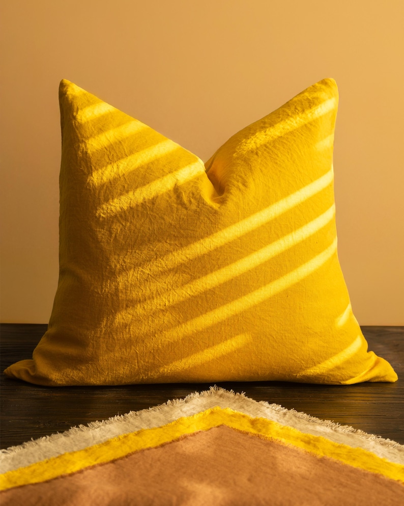 May include: A bright yellow decorative pillow with a textured surface. Diagonal shadows cast across the pillow create a striped pattern. A portion of a patterned blanket with yellow, white, and brown sections is visible below.