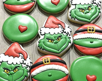 Könnte beinhalten: Auswahl an Weihnachtsplätzchen. Einige sind rund mit roter und grüner Glasur, die an den Gürtel und den Bauch des Weihnachtsmanns erinnern. Andere zeigen das Gesicht des Grinch mit einer Weihnachtsmütze. Einige Kekse haben eine grüne Basis mit einem roten Herzen.
