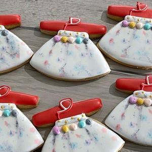 May include: Five sugar cookies decorated with red icing and white icing. The cookies are shaped like dresses with a red bodice and a white skirt. The skirts are decorated with a floral pattern and a row of colorful beads.