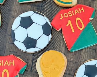 Puede incluir: Un conjunto de seis galletas de azúcar decoradas con un balón de fútbol, un trofeo y una camiseta con el nombre Josiah y el número 10. Las galletas están sobre una superficie de madera.