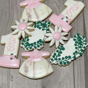 May include: A set of nine decorated sugar cookies featuring a pink and white floral theme. The cookies include two pink and white dresses, two white daisies with pink centers, two green wreaths, and two pink and white cookies with the word "one" written on them.