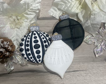Puede incluir: Tres galletas con forma de adorno navideño. Una es blanca con lunares negros, otra es negra con un diseño de cuadros y la tercera es blanca con un diseño de borlas. Cada una tiene una tapa plateada. Las flores de pascua blancas y una piña están en el fondo.