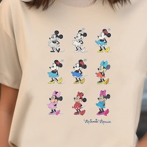 Puede incluir: Una camiseta beige con un estampado gráfico de Minnie Mouse. El gráfico presenta a Minnie Mouse en varias poses y atuendos, incluyendo un vestido rojo, un vestido azul y un vestido amarillo. El texto "Minnie Mouse" está impreso debajo del gráfico.