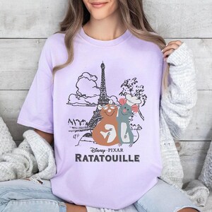 Può includere: Una maglietta color lavanda con una grafica di Remy e Linguini dal film Disney Pixar Ratatouille. La grafica presenta la Torre Eiffel, una nuvola e un albero. Il testo "Disney Pixar Ratatouille" è stampato sotto la grafica.