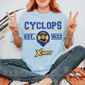 May include: Light blue t-shirt featuring the word "CYCLOPS" above a circular graphic of a superhero with the text "EST. 1832". Below the graphic is the word "X-MEN" in yellow. The shirt is a short-sleeved tee.