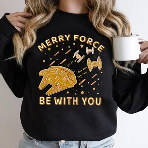 Puede incluir: Sudadera negra con un diseño de Star Wars con temática navideña. Presenta el texto "Merry Force Be With You" con naves espaciales estilo gingerbread y bastones de caramelo.