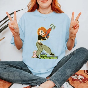 Puede incluir: Una camiseta azul claro con un gráfico de Kim Possible de la serie de dibujos animados de Disney. Kim sostiene un arma y está de pie junto a su compañero, Ron Stoppable. El texto "Disney's Kim Possible" está impreso debajo del gráfico.