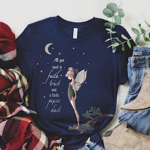Puede incluir: Una camiseta azul marino con un gráfico de Campanilla, un hada con alas, de pie en una rama. El texto "All you need is faith, trust and a little pixie dust" está impreso en la camiseta.