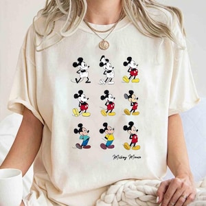 Puede incluir: Una camiseta blanca con un gráfico de Mickey Mouse en diferentes poses. El texto "Mickey Mouse" está impreso en la parte inferior de la camiseta.