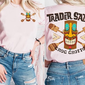May include: Light pink t-shirt featuring a colorful tiki design with crossed paddles on the front and back. The back of the shirt displays the text "Trader Sam's Frog Grotto" in brown lettering. The shirt is paired with blue denim shorts.