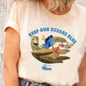 Può includere: Una maglietta bianca con una grafica di Dory, Nemo e Crush di Alla ricerca di Nemo. La grafica dice "Keep Our Oceans Blue" e ha il logo Disney Pixar Alla ricerca di Nemo.