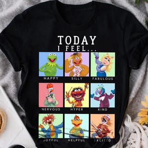 Op de afbeelding: Zwart T-shirt met een afbeelding van Sesamstraat-personages. De tekst "TODAY I FEEL..." staat bovenaan de afbeelding. Elk personage bevindt zich in een vierkant met een andere achtergrondkleur en een woord dat hun stemming beschrijft. De woorden zijn: HAPPY, SILLY, FABULOUS, NERVOUS, HYPER, KIND, JOYFUL, HELPFUL, EXCITED.