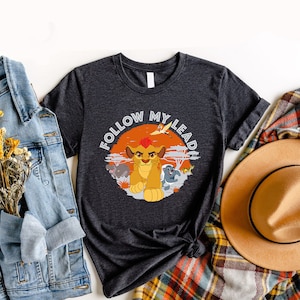 May include: A dark heather grey t-shirt with a graphic print of a lion cub from the Lion King movie. The graphic features the lion cub with a red mane, surrounded by other animals, and the text "Follow My Lead".