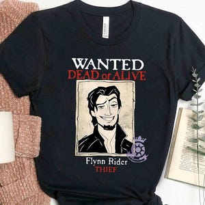 Puede incluir: Camiseta negra con un diseño de cartel de búsqueda con Flynn Rider de Enredados de Disney. El cartel dice "WANTED DEAD or ALIVE" y "Flynn Rider THIEF".