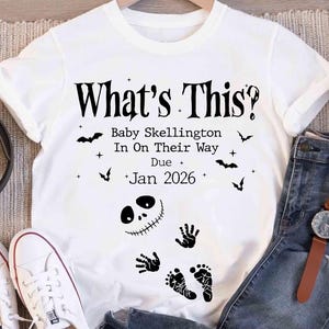A white t-shirt with a Halloween-themed design, featuring text and illustrations related to a baby named Skellington.