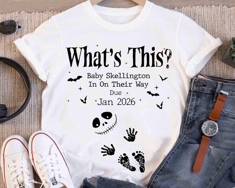 A white t-shirt with a Halloween-themed design, featuring text and illustrations related to a baby named Skellington.