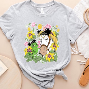 Puede incluir: Una camiseta gris con un diseño floral que presenta un caballo blanco con una diadema negra de orejas de Mickey Mouse y la palabra "Maximus" en ella. Un camaleón verde con una diadema negra de orejas de Mickey Mouse y la palabra "Pascal" en ella está al lado del caballo.