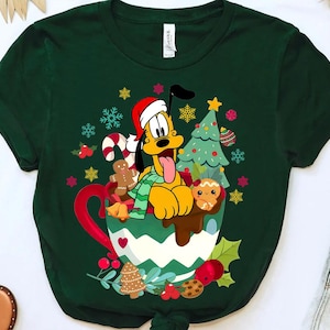 Peut inclure: T-shirt vert avec une image de dessin animé de Pluto le chien portant un chapeau de Père Noël et tenant une tasse de chocolat chaud. La tasse est décorée d'un sapin de Noël, de cannes en sucre et de biscuits au pain d'épices.