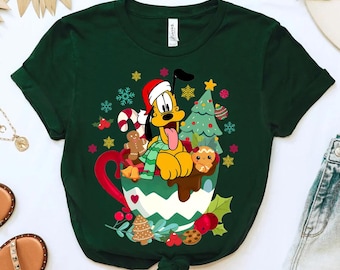 Puede incluir: Camiseta verde con una imagen de dibujos animados de Pluto el perro con un gorro de Papá Noel y sosteniendo una taza de chocolate caliente. La taza está decorada con un árbol de Navidad, bastones de caramelo y galletas de jengibre.