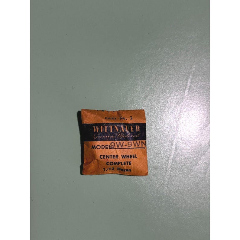 May include: A small, vintage brown paper package with black text and a black band. The text reads "WITTNAUER Genuine Material" and "MODEL 9W-9WN CENTER WHEEL COMPLETE 1/12 Dozen". The package is slightly crumpled.