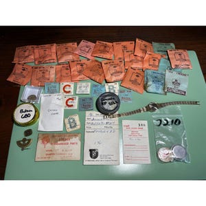 May include: A collection of vintage watch repair components including numerous small orange and blue paper envelopes labeled Bulova, a metal watch bracelet, a round container marked Bulova 680, a Caravelle RP 0162 tin, and various handwritten inventory cards.