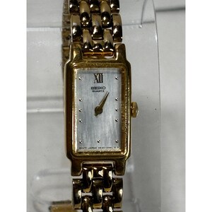 May include: A gold-tone Seiko Quartz wristwatch with a rectangular case and a silver-colored dial. The watch features a Roman numeral at the twelve o'clock position, stick hour markers, and a matching gold-tone metal link bracelet band.