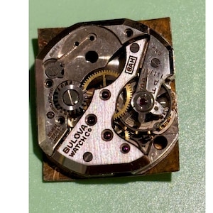May include: Close-up of a Bulova Watch Co. watch movement. The intricate mechanism features gears, jewels, and the text "Bulova Watch Co." The movement is set on a square, metallic base.