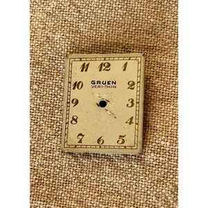 May include: A vintage Gruen Veri-Thin watch face with black numerals on a cream background. The square face has a small hole in the center and is surrounded by a thin, decorative border. The numbers 1 through 12 are clearly visible.