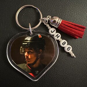 May include: Heart-shaped keychain featuring a hockey player in a black helmet. The keychain has a silver ring, a red tassel, and white beads spelling "JH 86". A personalized accessory for sports fans.