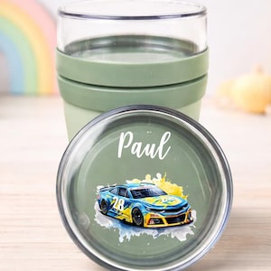 May include: A green and clear travel cup with a lid featuring a race car design and the name "Paul". The cup is part of a set, as indicated by the "Design 3" sticker. The background includes a teddy bear and children's toys.