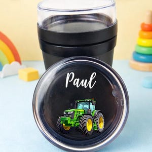 May include: Black travel mug with a clear lid, featuring a black lid with a green and yellow tractor design and the name "Paul" in white script. The mug is surrounded by toys, including a rainbow and a teddy bear.