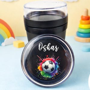 Könnte beinhalten: Schwarzer Reisebecher mit transparentem Deckel und Fußball-Design. Der Deckel trägt den Namen "Oskar" in weißer Schrift. Der Fußball ist von einem farbenfrohen Farbspritzer umgeben. Ein grüner Kreis mit "Design 1" ist ebenfalls sichtbar.