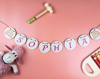 Peut inclure: Une bannière en bois rose et blanche avec le nom "Sophia" écrit en lettres roses. La bannière est enfilée sur une ficelle et comporte de petits cercles en bois avec des motifs arc-en-ciel à chaque extrémité. On voit également un mouton en peluche rose, un maillet en bois et un xylophone coloré.