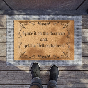 Puede incluir: Una alfombra de bienvenida marrón con un borde blanco y una inscripción negra que dice "Leave it on the doorstep and get the Hell outta here!" La alfombra está sobre un suelo de madera con un par de zapatos negros encima.