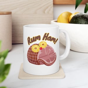 May include: White ceramic mug with the words "Rum Ham!" in brown script. The mug features a cartoon illustration of a ham, pineapple slices, and a cherry. The mug is sitting on a beige coaster.
