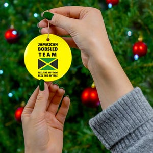 May include: A round, yellow Christmas ornament featuring the text "JAMAICAN BOBSLED TEAM" and the Jamaican flag. The ornament also includes the phrase "FEEL THE RHYTHM FEEL THE RHYME". The ornament is held against a background of a Christmas tree.