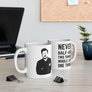 May include: Two white ceramic mugs. One mug features a black silhouette of a man with a mustache. The other mug has the text "NEVER HALF-ASS TWO THINGS WHOLE ASS ONE THING" in black.