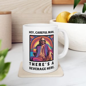 May include: White ceramic mug featuring a colourful illustration of a man holding a glass of beverage. The text on the mug reads "Hey, Careful Man, There's a Beverage Here."