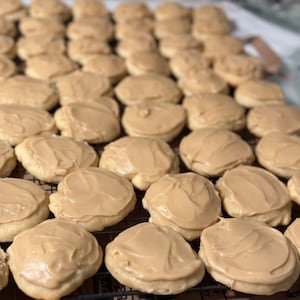 May include: A close-up of a wire rack filled with freshly baked sugar cookies, each topped with a generous layer of light brown icing.