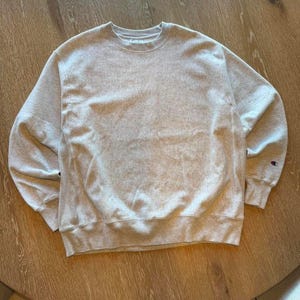 May include: A light grey Champion sweatshirt with a crew neck and long sleeves. The sweatshirt has a small Champion logo on the left sleeve cuff. The fabric appears to be a soft, comfortable material.