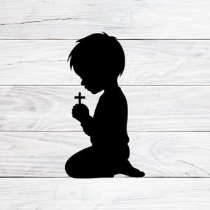 May include: A black silhouette of a child kneeling in prayer, holding a cross. The child's head is bowed, and the cross is held in front of them. The image is set against a white wooden background.