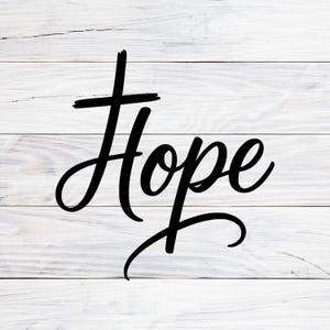 May include: Black cursive text that reads "Hope" on a white wooden plank background. The word is written in a flowing script with a cross above the letter "H". The background has a rustic, farmhouse style.
