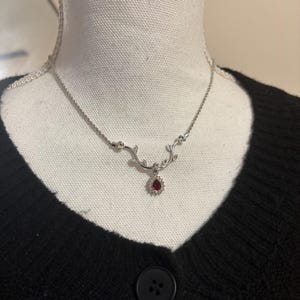 May include: A silver necklace with a delicate vine design. The necklace features small clear stones and a teardrop-shaped red gemstone pendant. The necklace is displayed on a white mannequin head, against a black sweater.