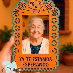 Puede incluir: Un marco de fotos naranja y turquesa con temática del Día de Muertos con la foto de una anciana sonriente. El marco presenta un diseño de calavera en la parte superior y el texto "YA TE ESTAMOS ESPERANDO" en la parte inferior.