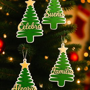 Puede incluir: Cuatro adornos verdes para árboles de Navidad con estrellas doradas y contornos blancos cuelgan de un árbol de Navidad. Cada adorno tiene una palabra dorada diferente: Celebra, Sueña, Familia y Alegria.