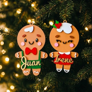 Puede incluir: Dos adornos navideños con temática de pan de jengibre. Uno tiene una pajarita roja y un peto verde con el nombre "Juan". El otro tiene un vestido rojo y el nombre "Irene". Ambos tienen detalles de glaseado blanco y están en un árbol de Navidad.