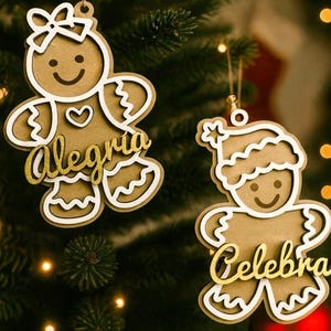 Puede incluir: Dos adornos navideños de hombre de jengibre. Un adorno tiene un lazo y la palabra "Alegria" en letras doradas. El otro adorno tiene una estrella y la palabra "Celebra" en letras doradas. Ambos cuelgan de un árbol de Navidad.